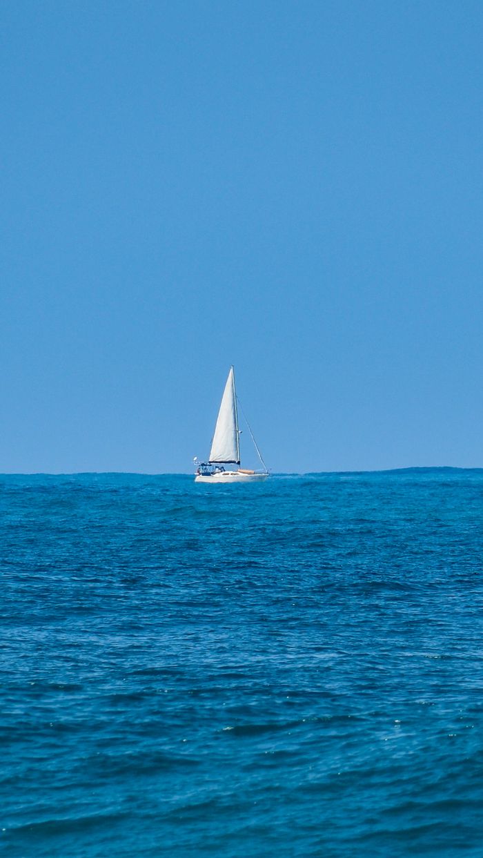 A solitary sailboat gracefully glides on the clear blue ocean under a bright summer sky.